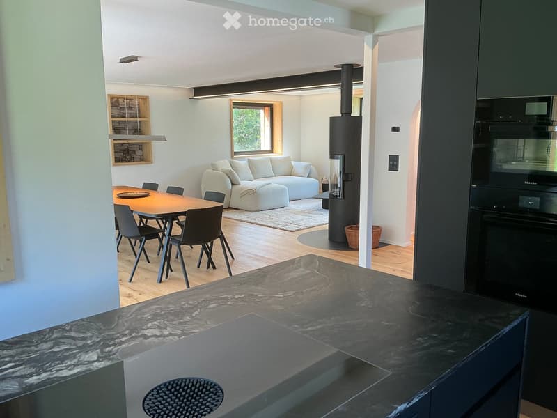 Modern open-plan living and dining area with a white sofa, ottoman, dining table with chairs, wood stove, and built-in oven.
