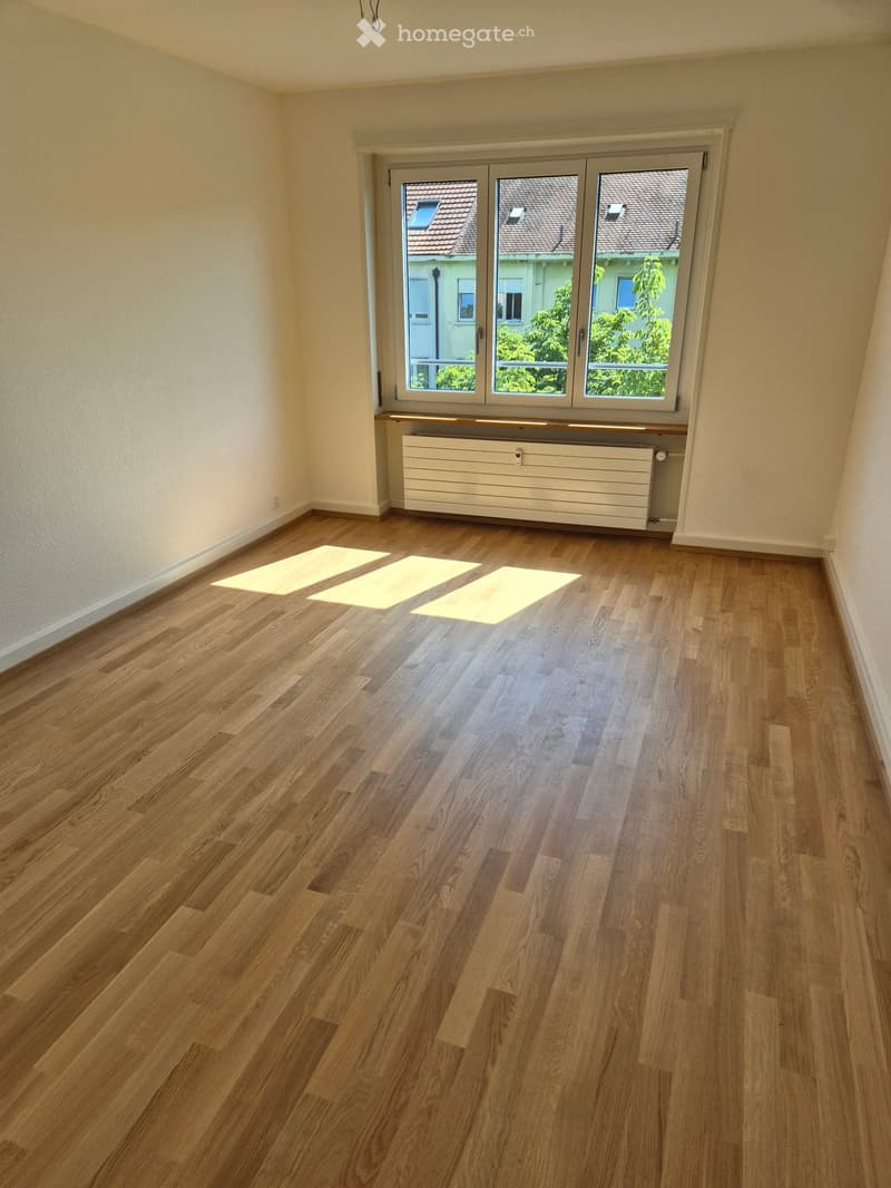 Empty room with wooden flooring, white walls, and a large window with a view of neighboring buildings and greenery. A radiator is visible below the window.