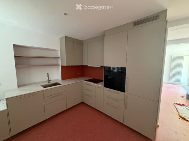 Modern kitchen with light-colored cabinets, a sink, an oven, and open shelving. The floor is a reddish-brown color, and the walls are white with a red accent wall behind the sink.