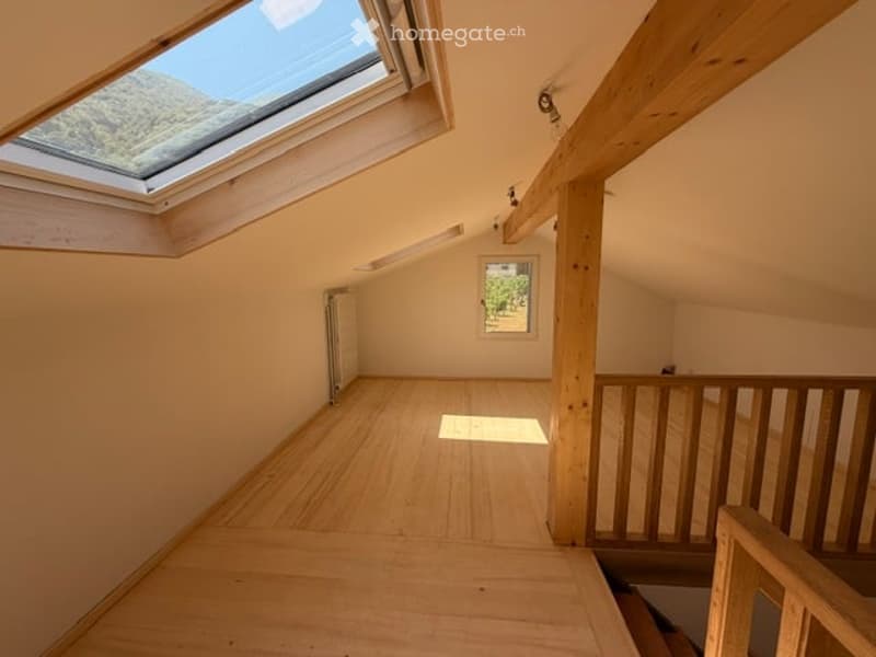 Empty attic room with wooden flooring, exposed wooden beams, skylight window, and a small window on the side wall.