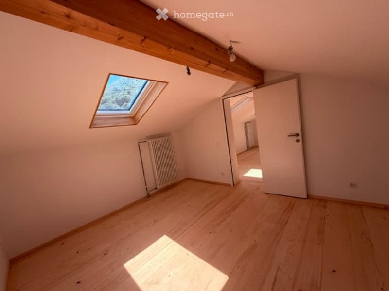 Empty attic room with wooden floor, exposed wooden beams, skylight window, radiator, and open doorway leading to another space.