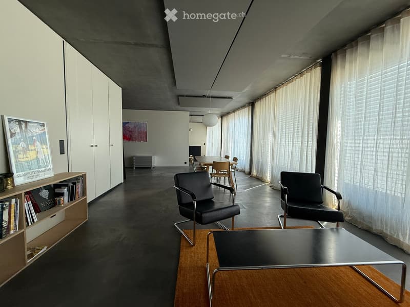 Modern, minimalist living space with dark chairs, a coffee table, a bookshelf, and large windows with white curtains.