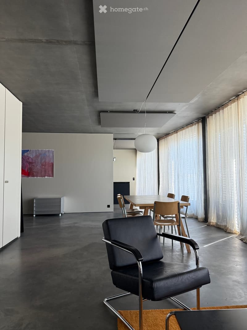 Modern, minimalist room with concrete floors and walls, featuring a black leather chair, a wooden dining table with chairs, large windows with white curtains, and a grey cabinet with a painting above it.