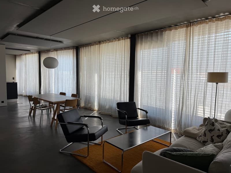 Modern living room with large windows covered by sheer curtains, a grey sofa with patterned cushions, a coffee table, two black armchairs, a dining table with chairs, and a floor lamp.