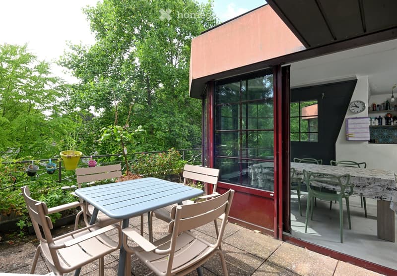 Balcony with outdoor furniture, including a table and chairs, surrounded by greenery and a view of the building interior.