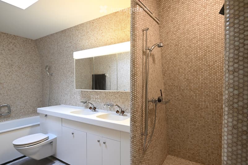 Modern bathroom with white cabinetry, double sink, toilet, bathtub, and separate shower area with a glass enclosure. The walls are covered in a textured, patterned tile.
