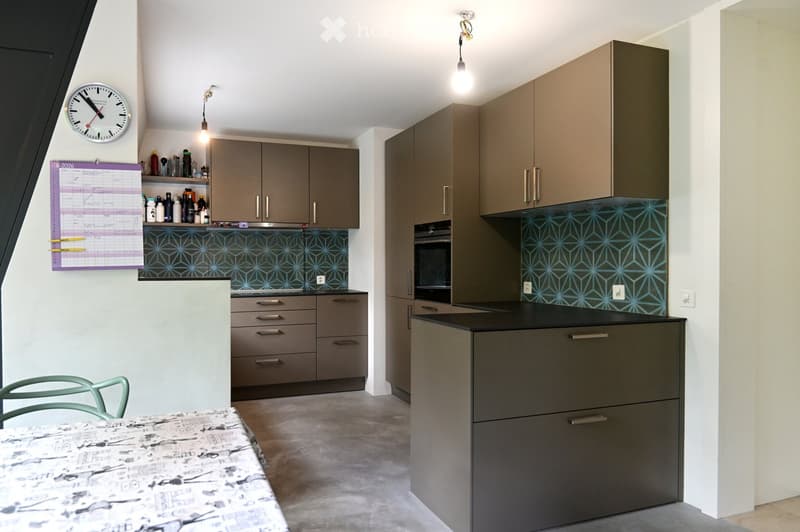 Modern kitchen with grey cabinets, a black countertop, and a geometric blue backsplash. A small dining area with a patterned tablecloth and a green chair is visible.