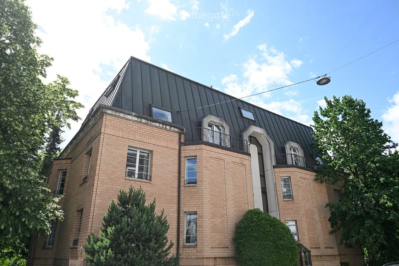 A multi-story brick building with a distinctive dark metal roof and several windows, surrounded by greenery and trees.