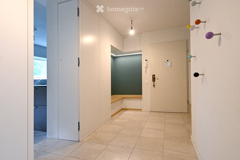 Clean, modern hallway with tiled flooring, white walls, and several doors. A small bench is visible in an adjacent room with a green accent wall.