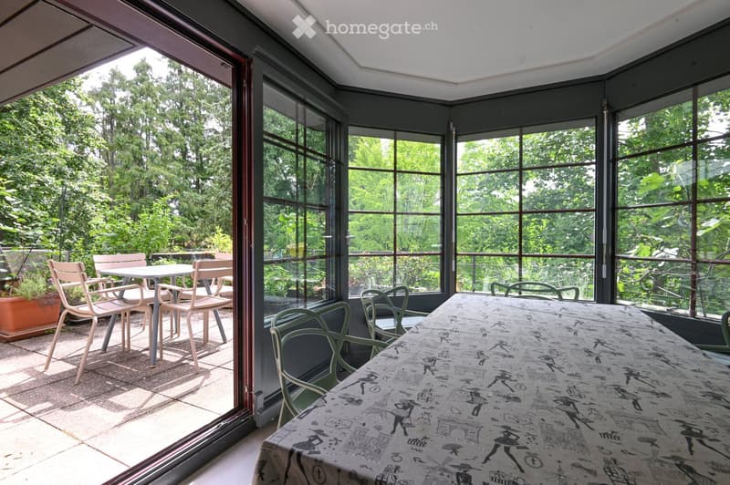 A sunlit room with large glass windows and a view of greenery, furnished with a long table covered in a patterned tablecloth and several chairs, both inside and outside the room.