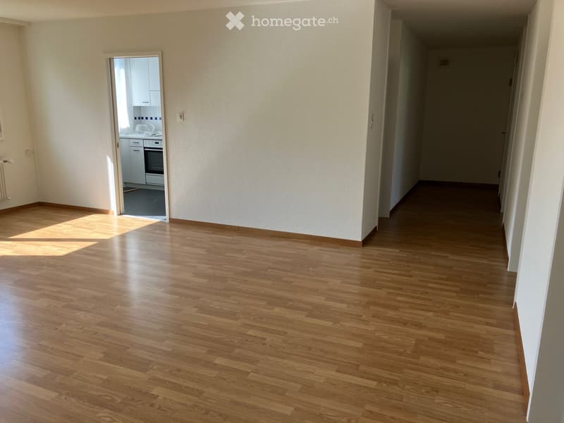 Empty room with wooden flooring and white walls, featuring a doorway leading to a kitchen area.