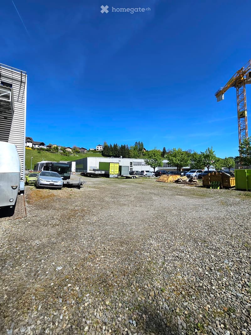 Gravel parking lot with several vehicles, including cars, a bus, and a trailer, surrounded by trees and buildings under a clear blue sky.