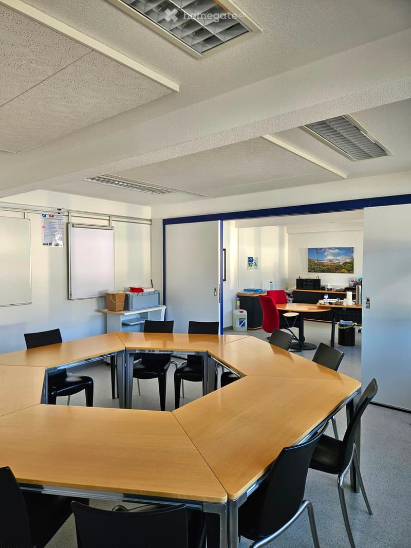 A modern conference room with a large, curved wooden table surrounded by black chairs, a smaller table with red chairs, and a whiteboard on the wall.