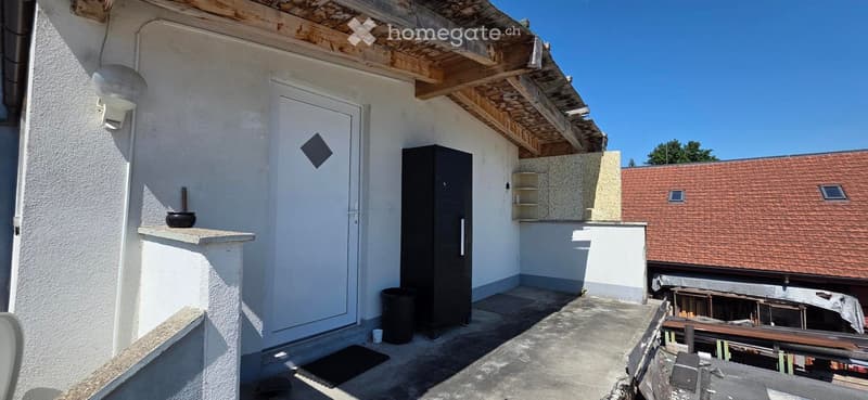 Balcony with white walls, a door, a small shelf, and a black cabinet. The roof is partially exposed with wooden beams.