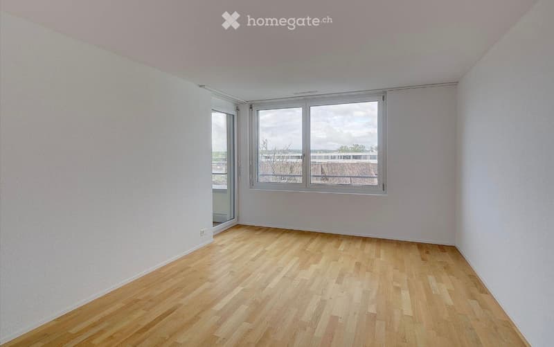 Empty room with light wooden flooring, white walls, and large windows offering a view of the outside area.