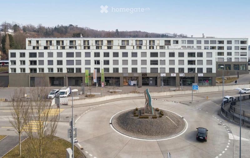 Modern multi-story building with numerous windows and a parking area in front, featuring a central sculpture and several parked vehicles.