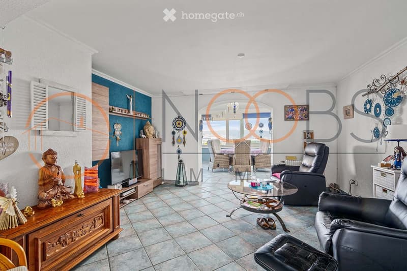 A modern living room with a tiled floor, white walls, and a mix of contemporary and decorative furniture. Features include a black leather sofa, a glass coffee table, a wooden cabinet with a TV, and a dining area visible in the background.