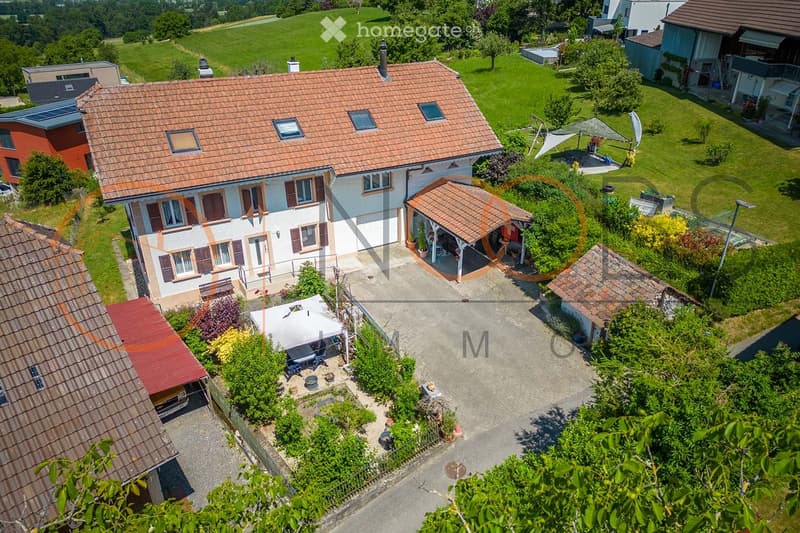 Aerial view of a two-story house with a red-tiled roof, surrounded by a well-maintained garden and driveway. The house features multiple windows, a covered patio area, and a small outdoor seating area with a table and chairs.