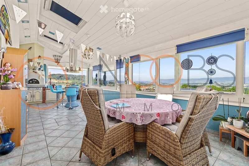 Bright, airy dining and kitchen area with large windows offering scenic views. Features a round dining table with wicker chairs, a kitchen island with bar stools, and decorative elements including vases and a chandelier.