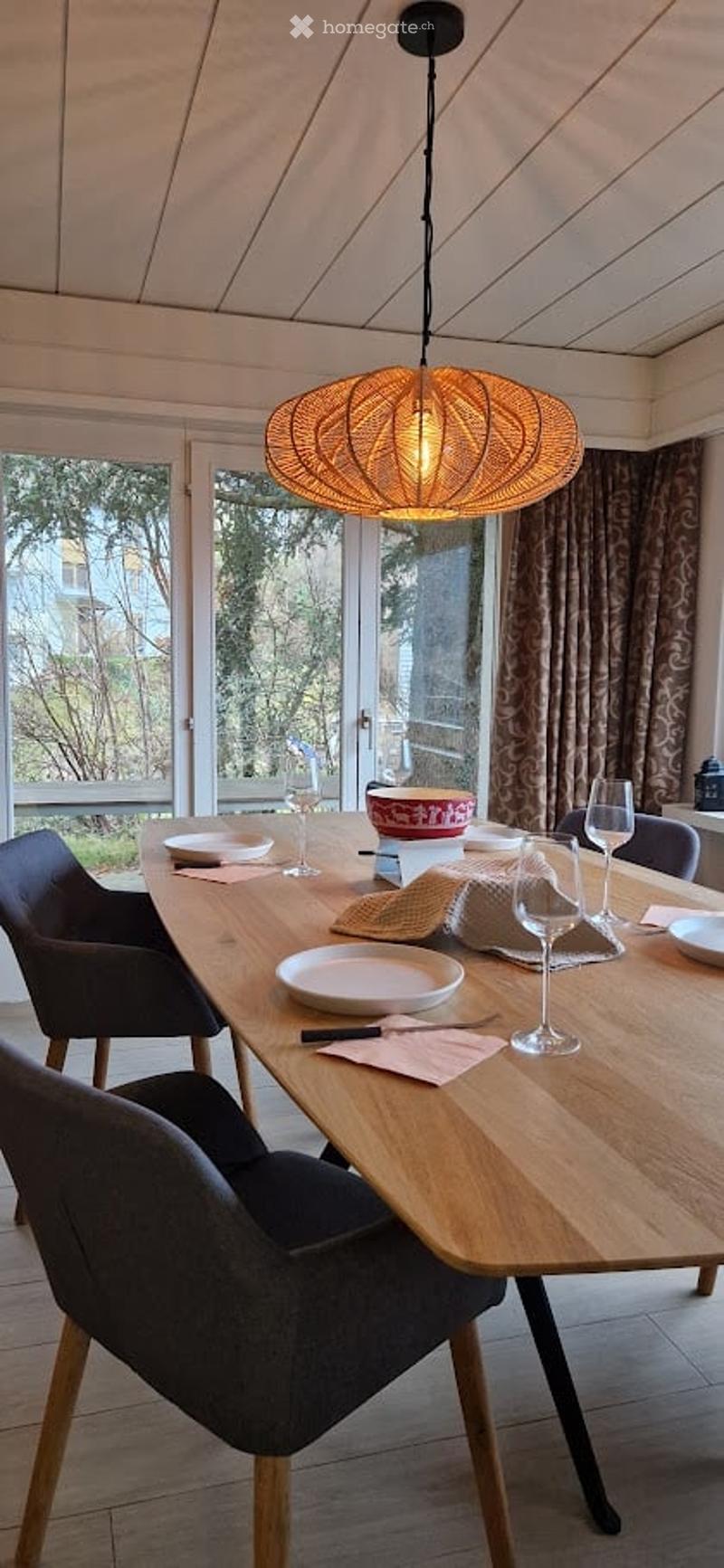 Dining area with a wooden table, chairs, wine glasses, plates, and a decorative bowl. Large windows with curtains and a hanging pendant light.