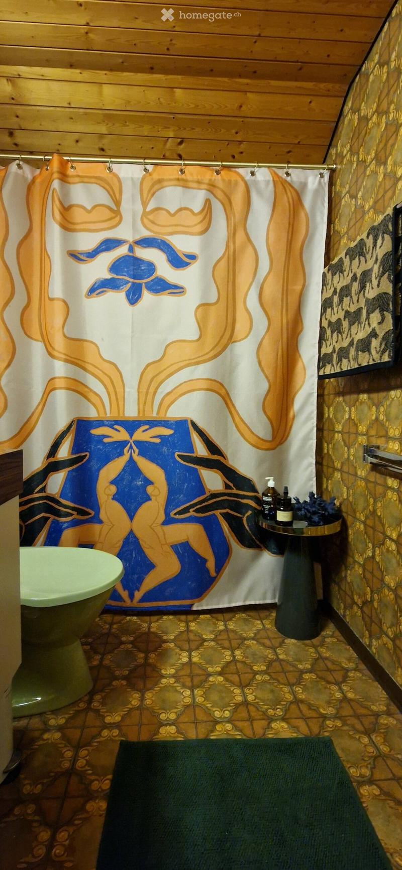 Bathroom with a green toilet, a small round table with bottles, a colorful shower curtain, and a patterned tile floor.