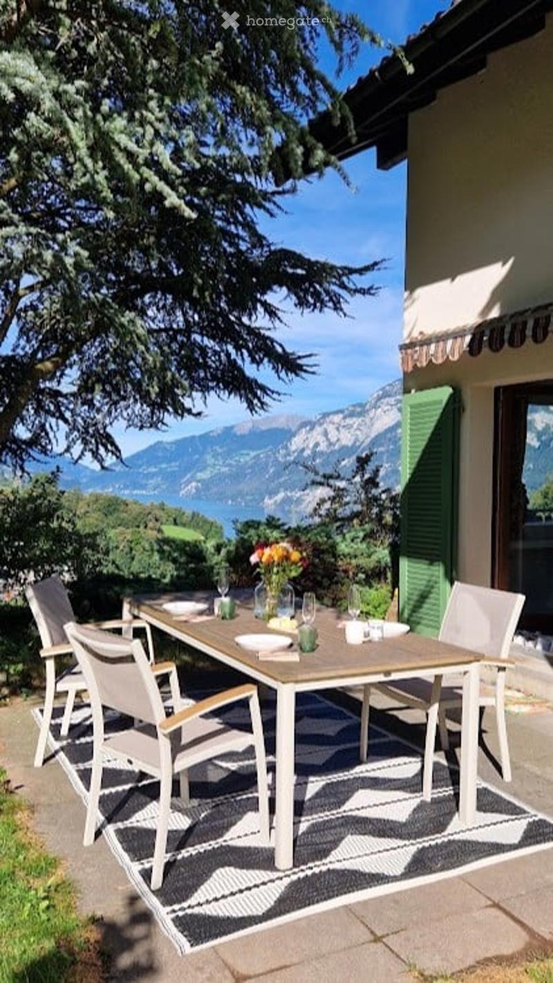 Outdoor dining area with a wooden table, white chairs, and a flower arrangement, overlooking a scenic mountain and lake view.