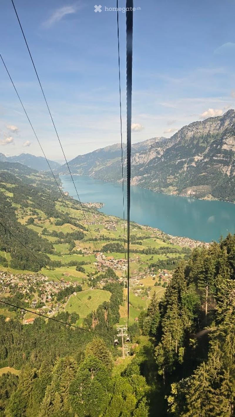 Panoramic view from a cable car showing a town nestled in green hills, a lake, and surrounding mountains under a clear blue sky.