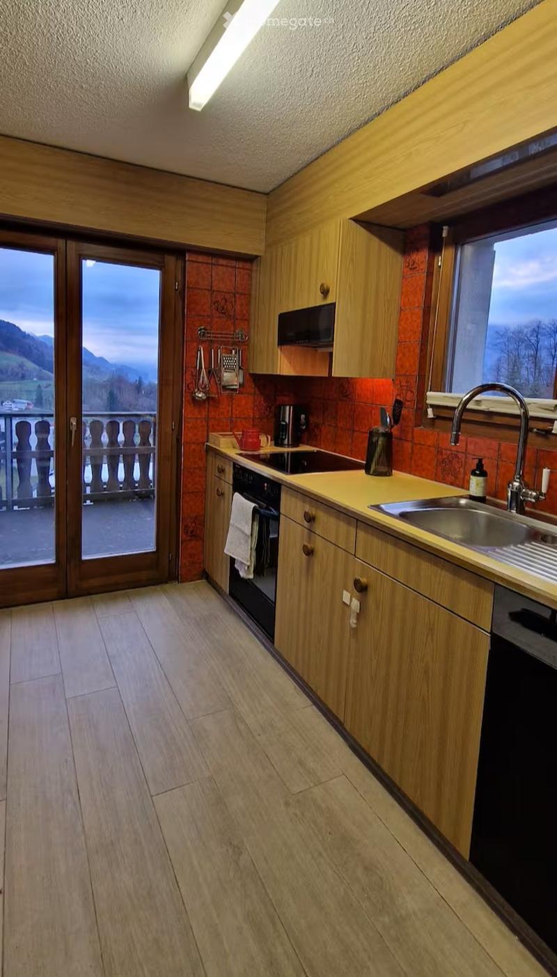 Modern kitchen with wooden cabinets, light-colored countertops, and a view of the mountains through large windows. Includes a sink, oven, microwave, coffee maker, and a towel hanging on the oven door.