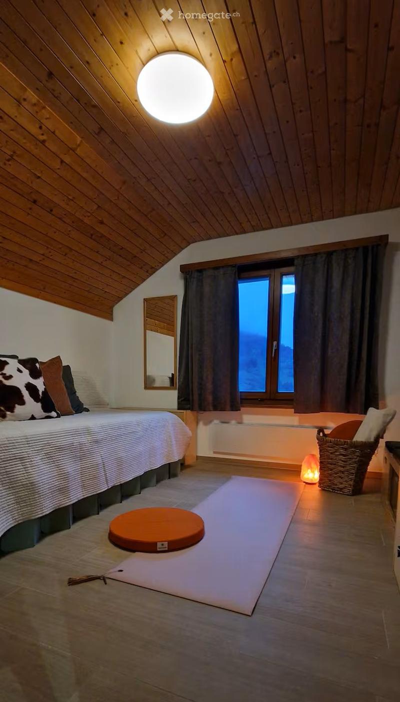 Cozy bedroom with wooden slanted ceilings, a large bed with patterned pillows, a meditation mat, a round cushion, a wicker basket with a small lamp, and a large window with dark curtains.