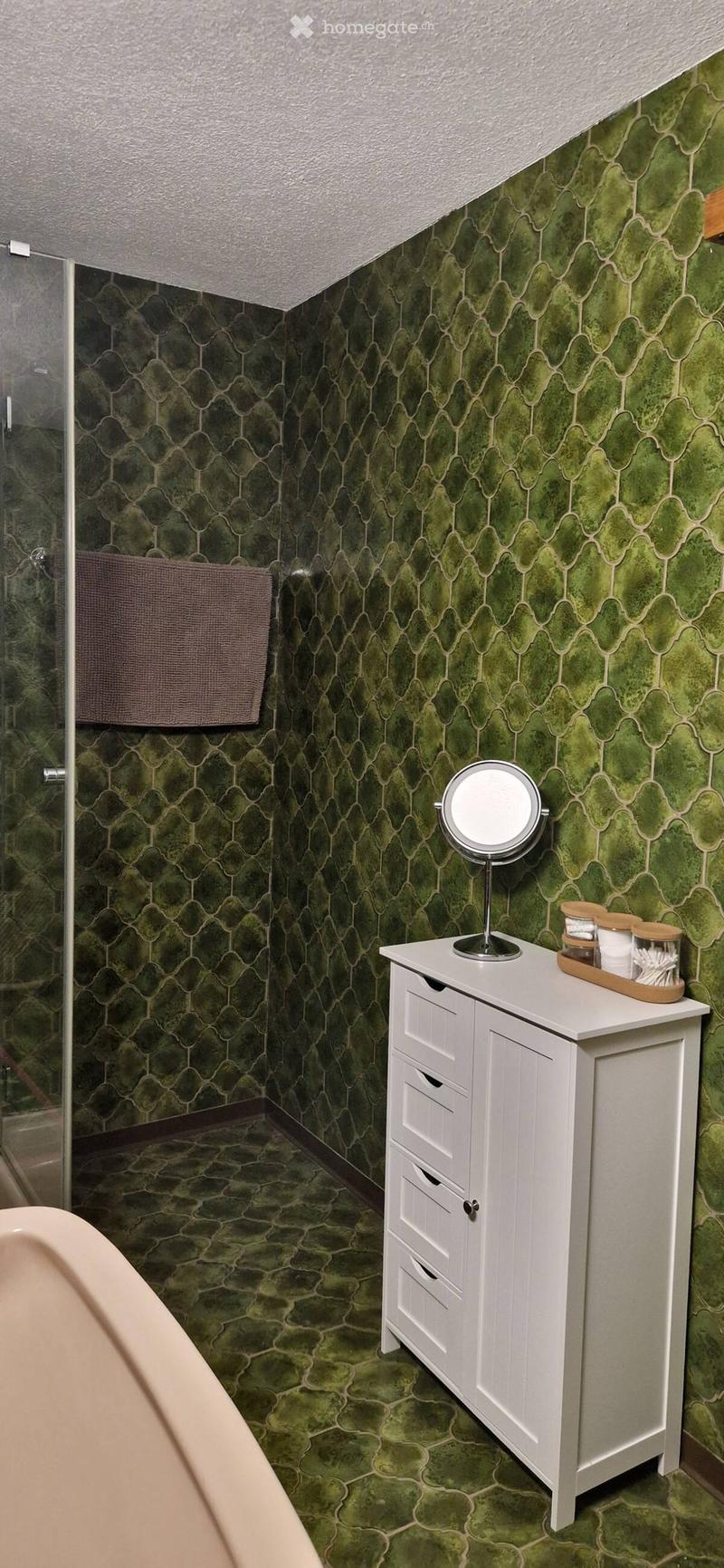 Bathroom with green patterned tiles, white vanity with drawers, round mirror, and a towel hanging on the wall.