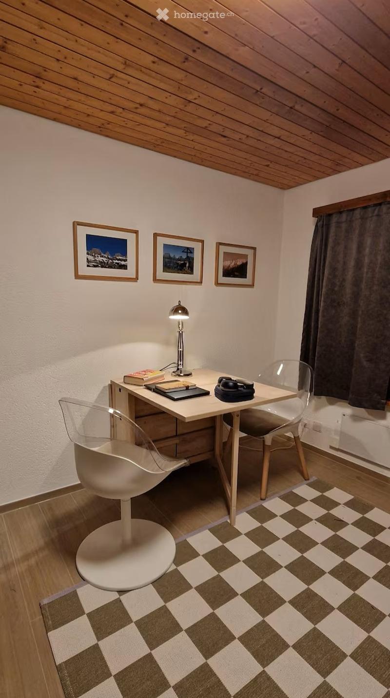 A small, cozy room with wooden walls and ceiling, featuring a modern desk with a lamp, books, and a camera, accompanied by two contemporary chairs and a checkered rug.