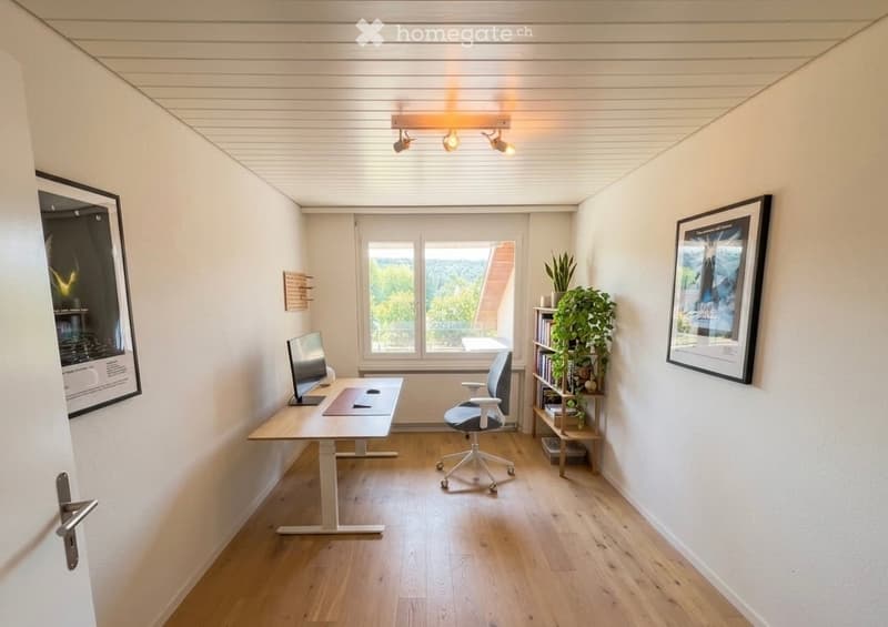 Modern, minimalist office space with light wooden flooring, white walls, and large windows. Includes a standing desk, ergonomic chair, computer screen, bookshelf with books and plants, and framed artwork on the walls.