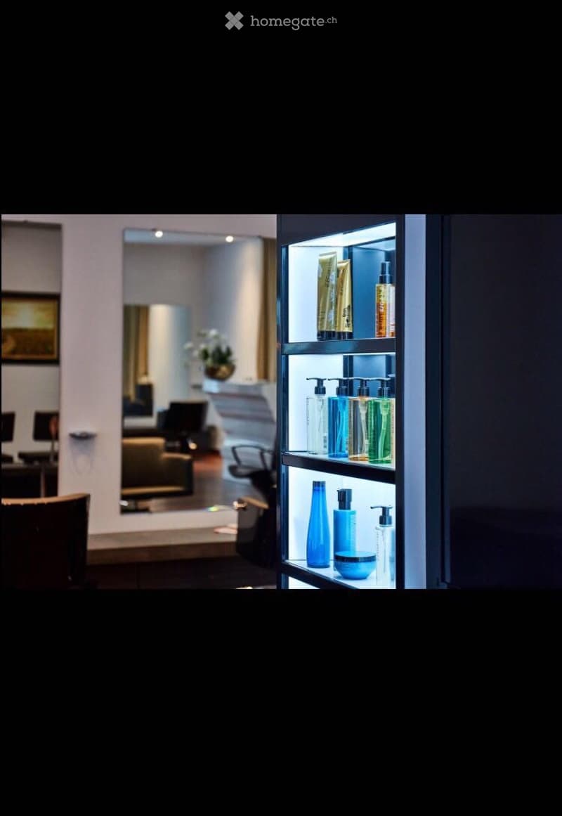 A modern, well-lit room with a sleek, illuminated shelf displaying various bottles and containers. The background shows a cozy seating area with a sofa, chair, and a small table, complemented by a mirror and a flower vase on a side table.