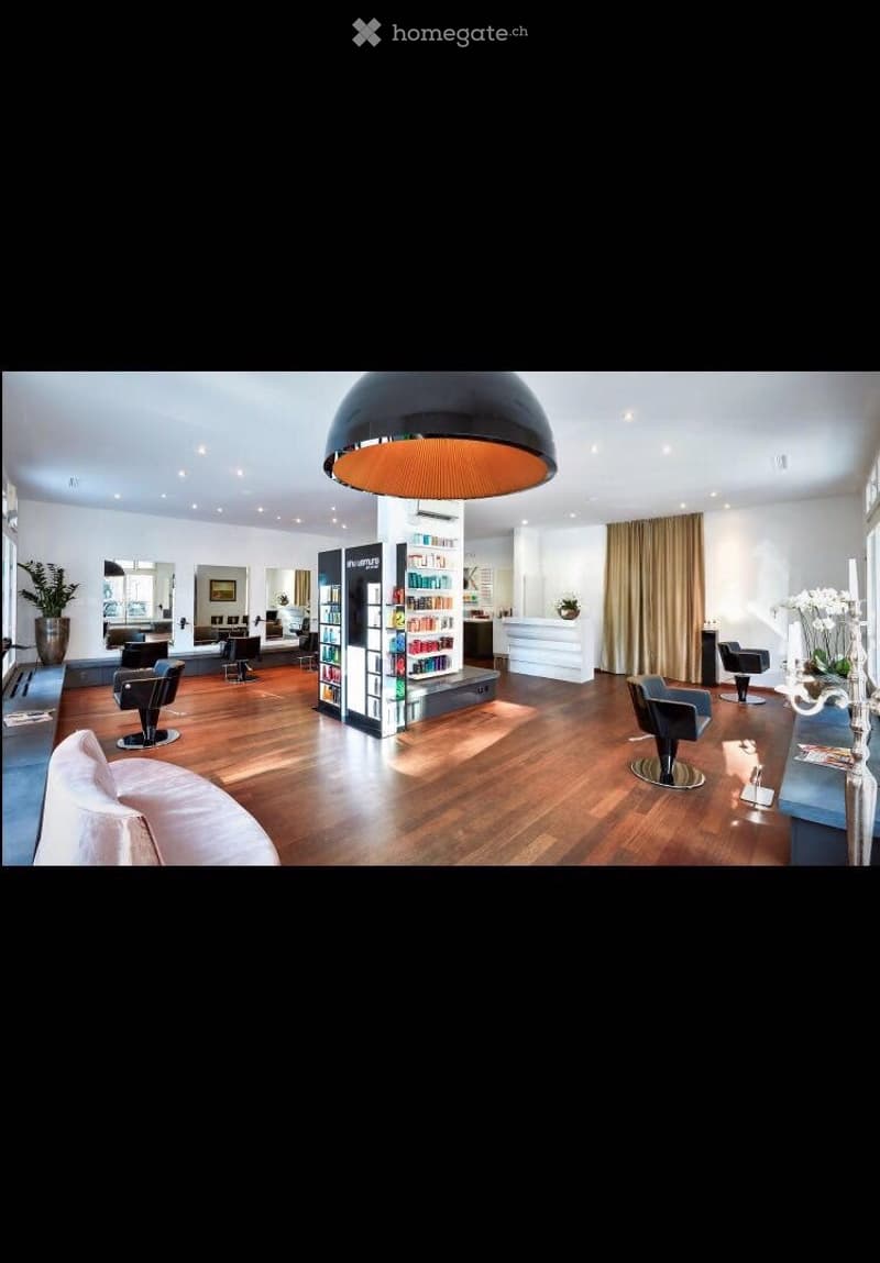 Modern, spacious salon with wooden flooring, large round pendant light, several chairs, a sofa, shelves with products, and a reception desk.