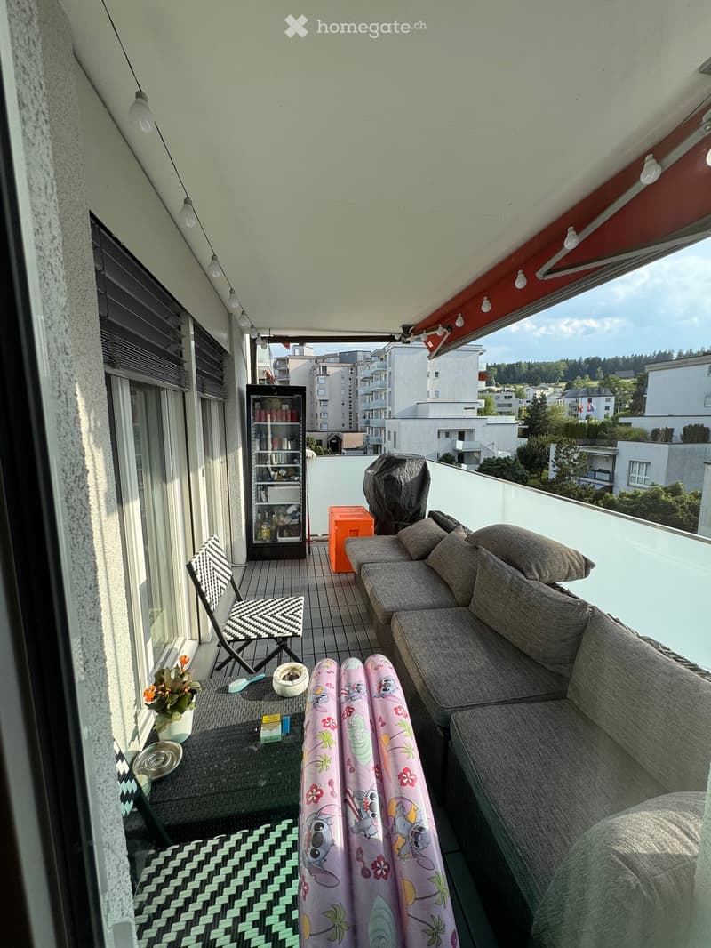 Balcony with a grey sofa, small table, folding chair, refrigerator, and a colorful blanket draped over the sofa.