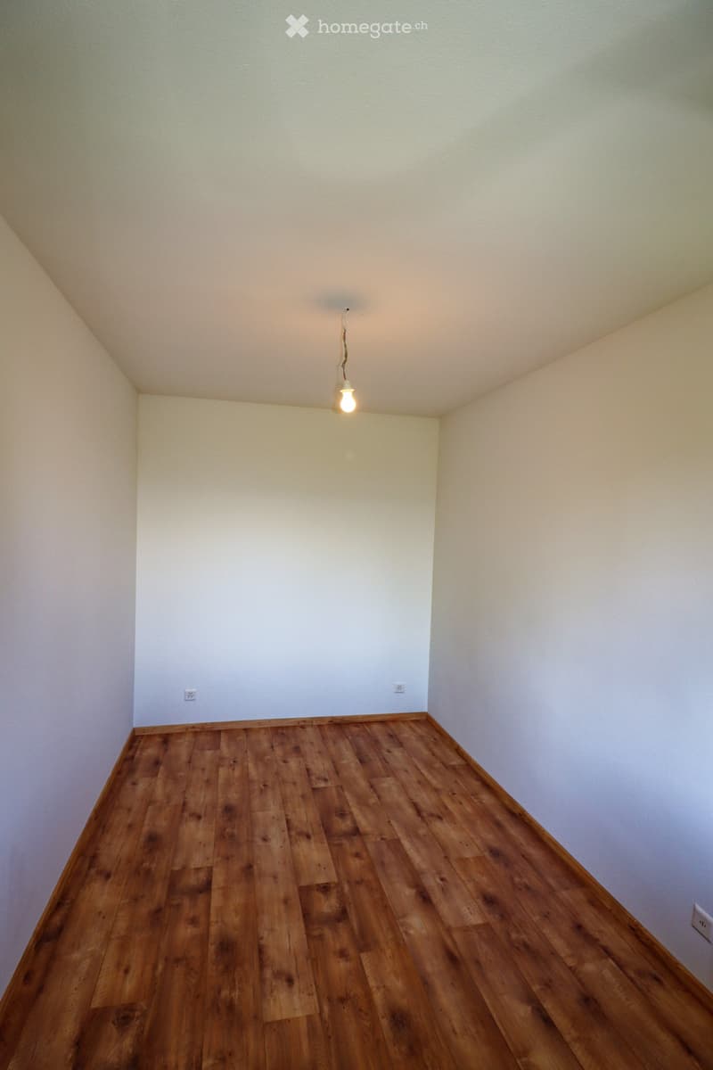 Empty room with wooden flooring and white walls, featuring a single hanging light fixture and electrical outlets.