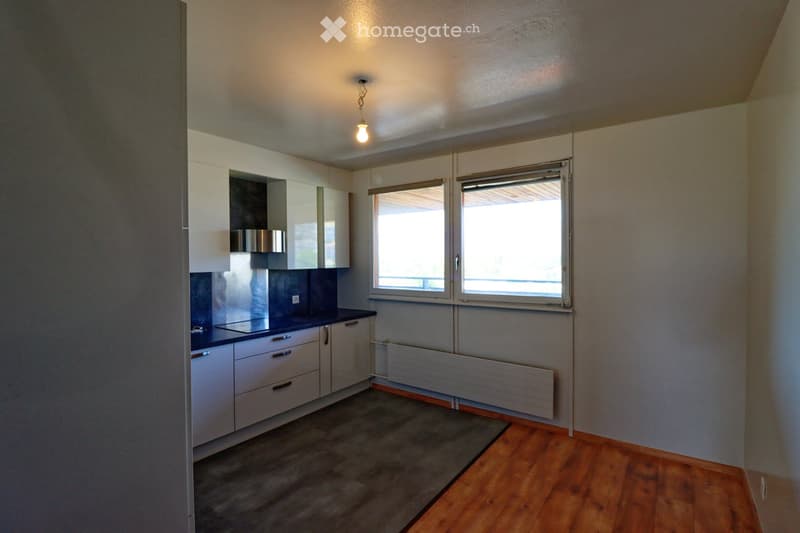 Modern kitchen with white cabinets, dark countertop, and wooden flooring. Features a stove, radiator, and large windows with blinds.