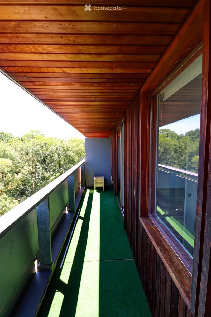 Wooden balcony with green carpeting, large windows, and a small yellow stool at the end.