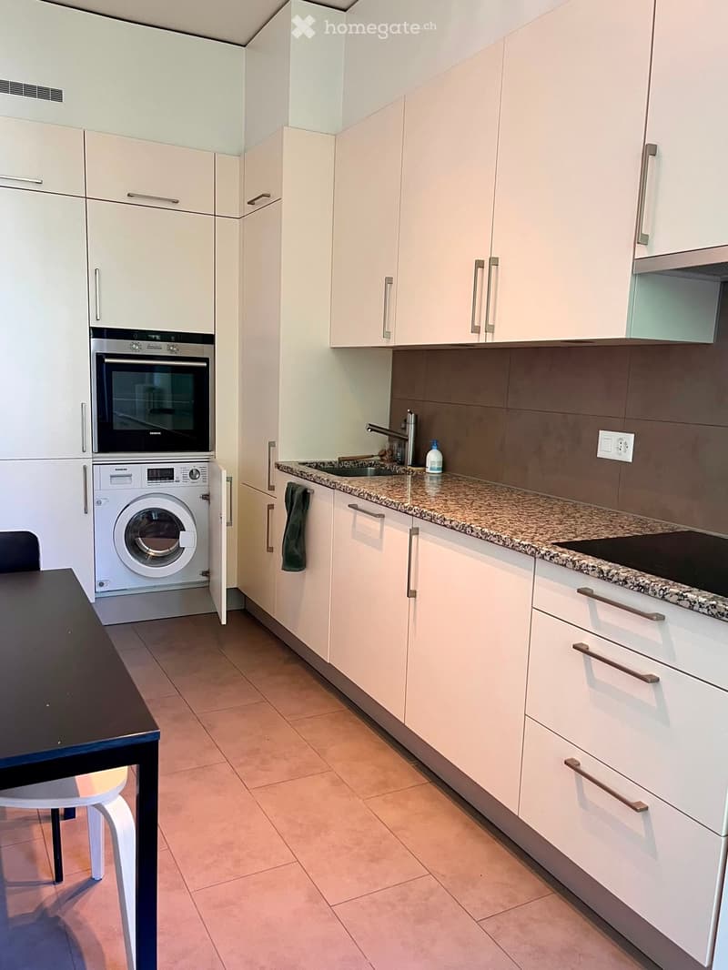 Modern kitchen with white cabinets, granite countertop, built-in oven, washing machine, and a small dining area with a table and chair.