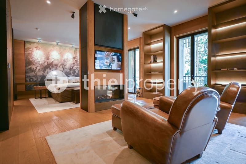 Modern living room with wooden flooring, large windows, leather armchairs, sofa, coffee table, fireplace, and built-in shelving with books and decorative items.