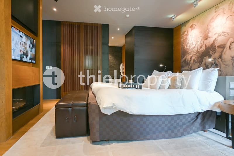 Modern bedroom with a large bed, nightstand, leather stool, fireplace, and a mounted screen.
