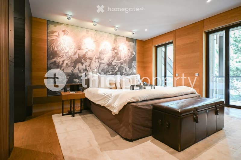 Modern bedroom with a large bed, nightstand, leather stool, and a large artwork on the wall. The room features wooden flooring and large windows with a view.