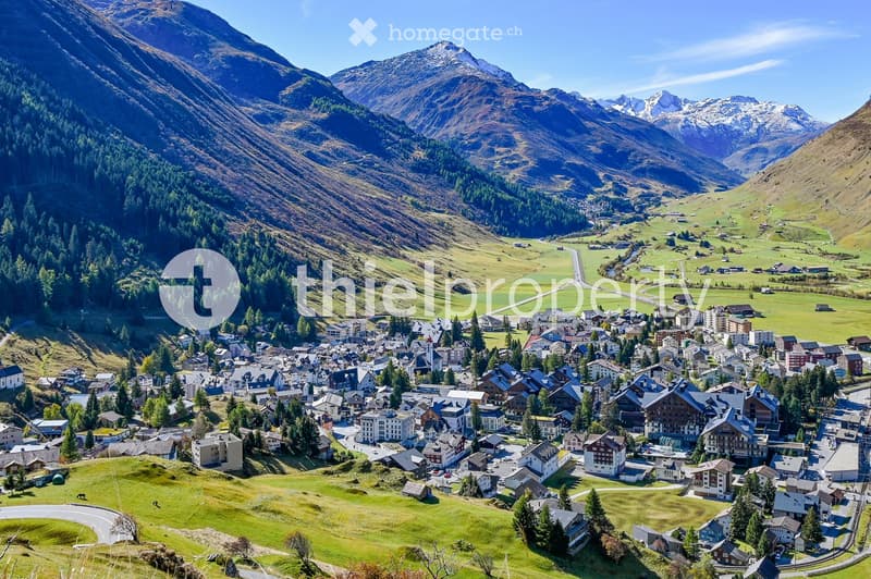 A picturesque village nestled in a lush green valley surrounded by towering mountains. The village features numerous houses and buildings with varied architectural styles, surrounded by dense forests and open green fields.