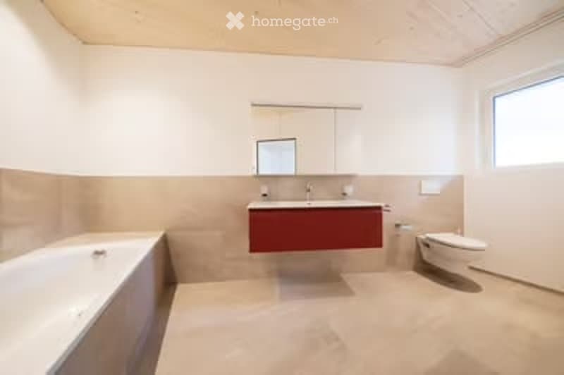 Modern bathroom with a white bathtub, red vanity, and a toilet. The walls are painted in a light beige color, and the floor is tiled in a light beige color as well. There is a large window on the right side of the room, allowing natural light to enter.
