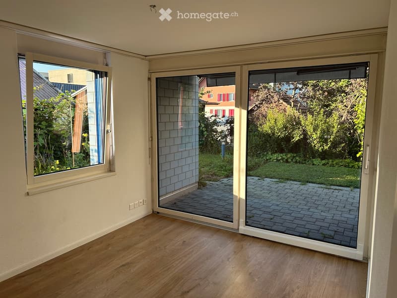 Empty room with wooden flooring, large sliding glass doors leading to a paved patio and garden area, and a window with a view of neighboring houses and greenery.