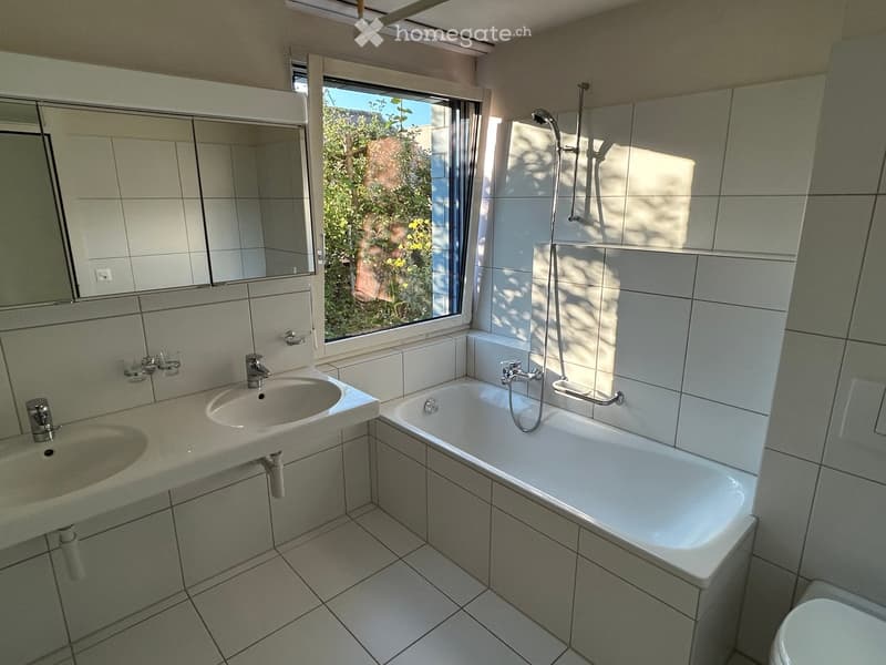 Modern bathroom with white tiled walls and floor, featuring a double sink, bathtub with shower, and a large mirror above the sink. The room has a window with a view of greenery outside.