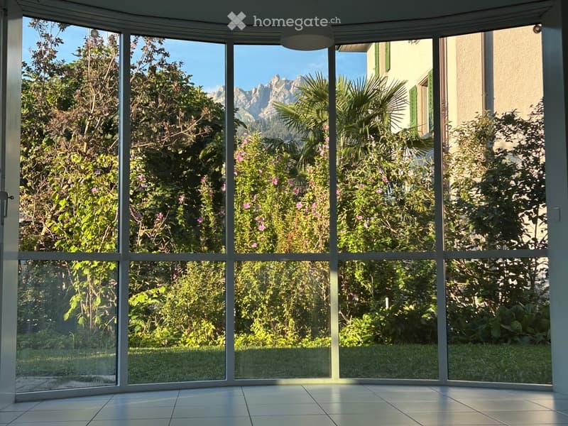 Empty room with large glass windows offering a view of a garden and mountains. The floor is tiled, and the space is well-lit with natural light.