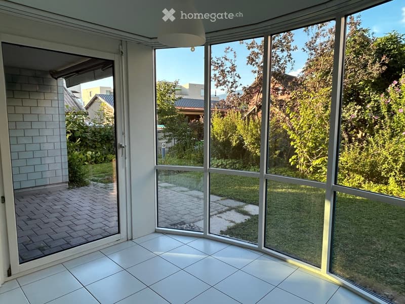 Bright, empty room with large glass windows and sliding doors leading to a garden area. The floor is tiled, and the walls are white.