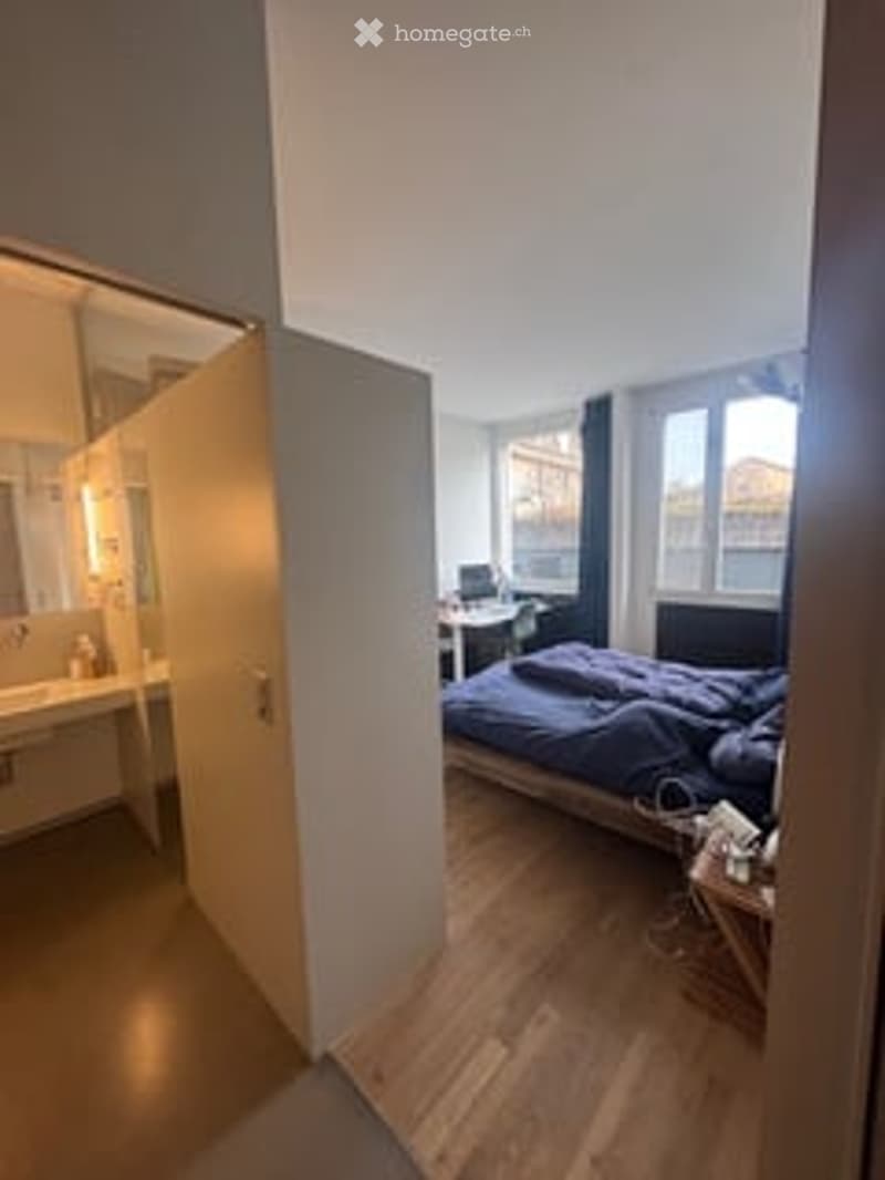 Cozy bedroom with wooden flooring, a large bed with blue bedding, a nightstand, a mirror, a sink, and a desk with a chair.