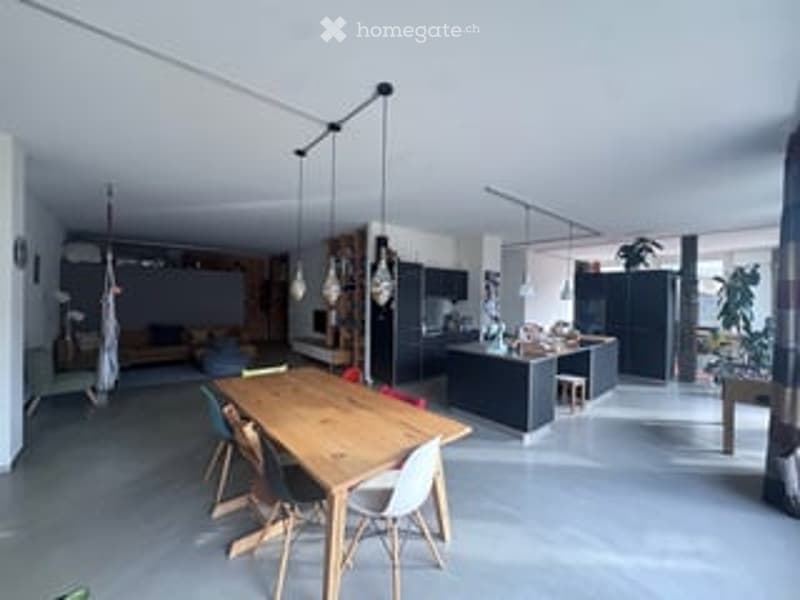 Open-plan living and dining area with modern kitchen, wooden dining table, pendant lights, and a couch in the background.